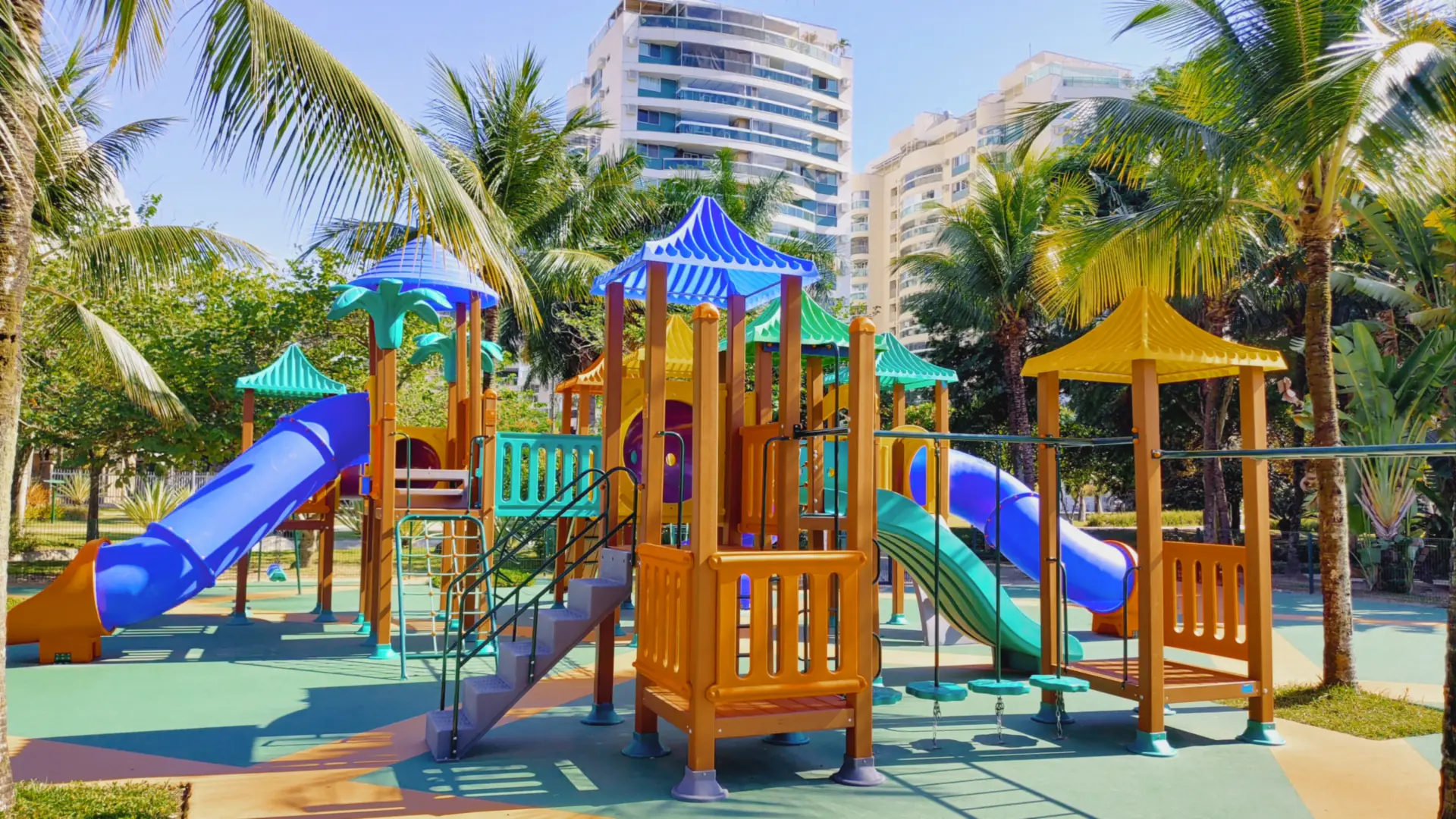Playground grande com torres de telhados coloridos e escorregadores em tubo, entre palmeiras e prédios, na Associação Rio 2, Barra Olímpica