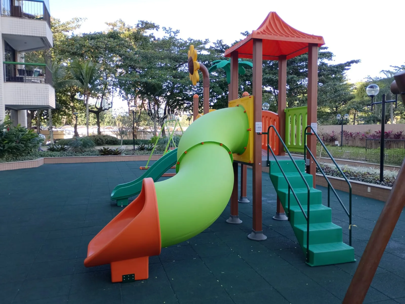 Playground com torre, telhado e tubo de escorregador sobre piso emborrachado, entre o jardim e o prédio do condomínio